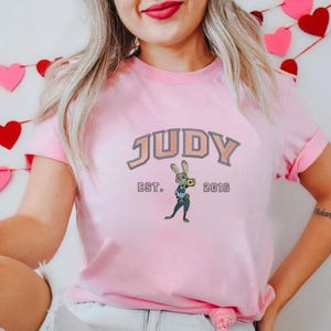 May include: Pink t-shirt featuring the name "JUDY" in a peach-colored, outlined font. Below the name is "EST. 2016" and a cartoon image of a rabbit character. The shirt has short sleeves.