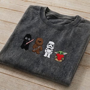 May include: Charcoal grey t-shirt with embroidered Star Wars characters: Darth Vader, Chewbacca, a Stormtrooper, and Grogu holding a heart. The shirt has a crew neck and a faded look.