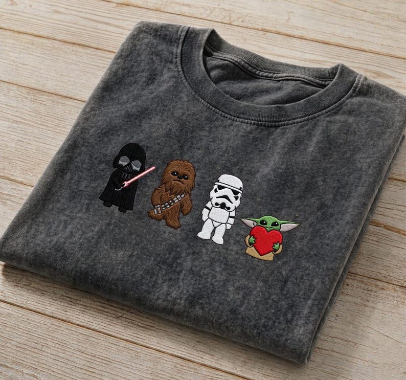 May include: Charcoal grey t-shirt with embroidered Star Wars characters: Darth Vader, Chewbacca, a Stormtrooper, and Grogu holding a heart. The shirt has a crew neck and a faded look.
