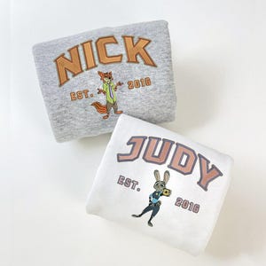 May include: Two folded sweatshirts, one gray and one white. The gray sweatshirt has the name "NICK" in orange lettering with a fox graphic and "EST. 2016". The white sweatshirt has "JUDY" in orange lettering with a bunny graphic and "EST. 2016".