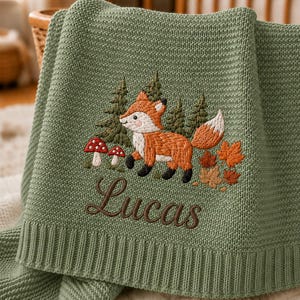Könnte beinhalten: Eine salbeigrüne Strickdecke mit einer Stickerei, die einen Fuchs, Tannenbäume, rote Pilze und Herbstblätter zeigt. Der Name Lucas ist in brauner Schreibschrift unter dem Fuchs eingestickt. Die Decke hat ein strukturiertes Strickmuster.