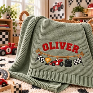 May include: A sage green knitted blanket featuring an embroidered design of a rope with a chequered flag, a gold trophy, a red race car with the number one, a stack of black tyres, and another chequered flag, with the name OLIVER in red.