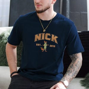 May include: Navy blue t-shirt with the name "NICK" in a brown arched font, and a cartoon fox character wearing a green tie. The text "EST. 2016" is below the fox. The shirt is short-sleeved.