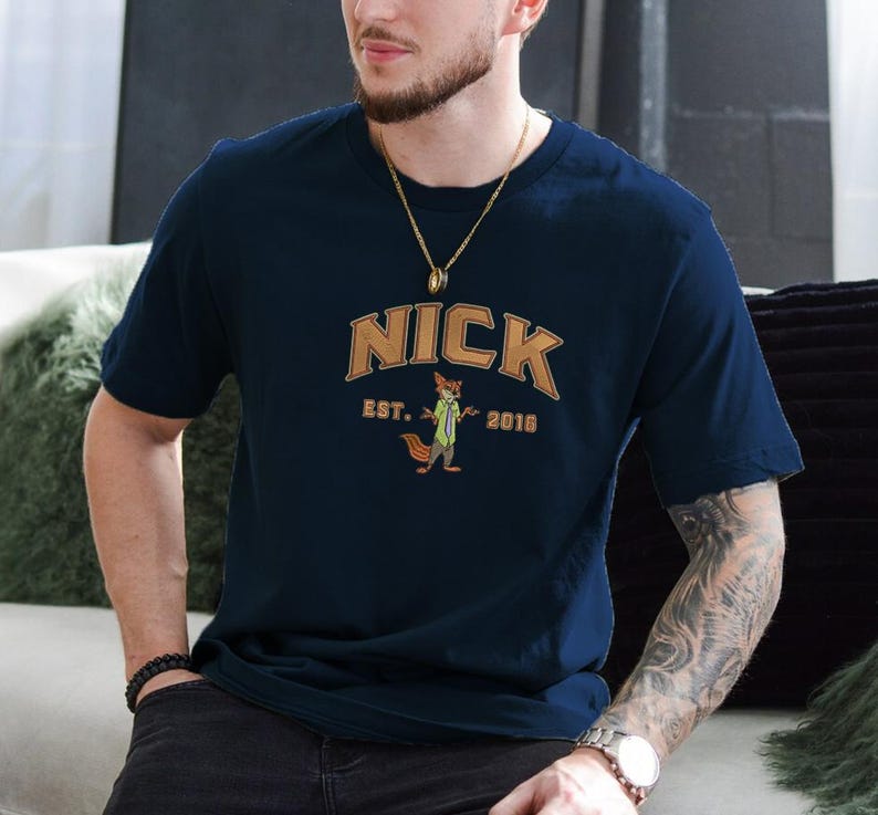 May include: Navy blue t-shirt with the name "NICK" in a brown arched font, and a cartoon fox character wearing a green tie. The text "EST. 2016" is below the fox. The shirt is short-sleeved.