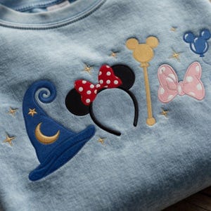 Pode incluir: Camisola azul clara com desenhos bordados da Disney. Apresenta um chap&eacute;u de feiticeiro azul com uma lua crescente, orelhas da Minnie com um la&ccedil;o vermelho de bolinhas, uma varinha do Mickey Mouse, um la&ccedil;o rosa de bolinhas e estrelas.
