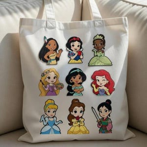 Könnte beinhalten: Weißer Canvas-Tote-Bag mit farbenfrohen Cartoon-Illustrationen verschiedener Prinzessinnen. Die Prinzessinnen sind in ihren ikonischen Outfits und Posen dargestellt. Die Tasche hat ein schlichtes Design mit rechteckiger Form und zwei Griffen.