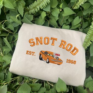 Puede incluir: Sudadera beige con el texto bordado "SNOT ROD EST. 1968" sobre un coche de dibujos animados naranja. La sudadera se muestra sobre un lecho de hojas verdes.