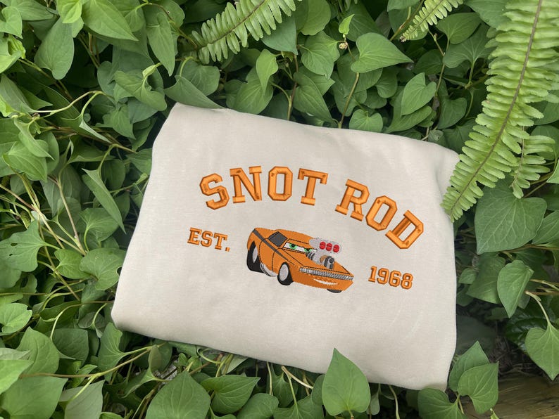 Puede incluir: Sudadera beige con el texto bordado "SNOT ROD EST. 1968" sobre un coche de dibujos animados naranja. La sudadera se muestra sobre un lecho de hojas verdes.