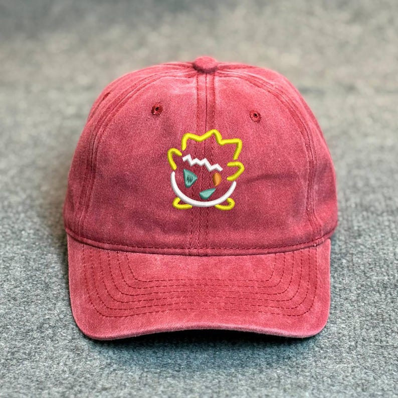 May include: A maroon baseball cap with a curved brim. The cap features an embroidered design of a cartoon character in yellow, white, and green. The cap is made of a soft, textured fabric and has a classic six-panel design.