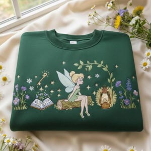 May include: A dark green sweatshirt featuring an embroidered fairy scene. The design includes a fairy, key, open book, lantern, and flowers. The sweatshirt is made of a soft material and is a crew neck style.