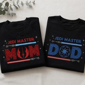 May include: Two black t-shirts with the words "JEDI MASTER" above the words "MOM" and "DAD" in red and blue, respectively. The "MOM" shirt features a red symbol, while the "DAD" shirt has a blue symbol. Both shirts have lightsaber designs.