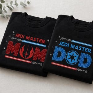 May include: Two black t-shirts with "JEDI MASTER" and lightsaber designs. One shirt says "MOM" in red glitter, the other says "DAD" in blue glitter with a circular emblem. A fun gift for Star Wars fans.