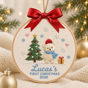 May include: A circular wooden embroidery hoop ornament featuring a cream fabric background with a stitched design of a teddy bear wearing a red hat and blue scarf, a green Christmas tree, a gift box, snowflakes, and hearts. The text reads "Luca's FIRST CHRISTMAS 2026" in blue, topped with a red satin ribbon bow.