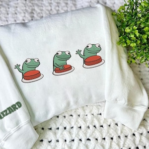 Könnte beinhalten: Weißes Sweatshirt mit einem Cartoon-Frosch-Design. Drei grüne Frösche sind auf roten Knöpfen abgebildet. Das Wort "LIZARD" ist in Grün auf dem linken Ärmel aufgedruckt. Das Sweatshirt ist auf einer weißen Oberfläche gefaltet.