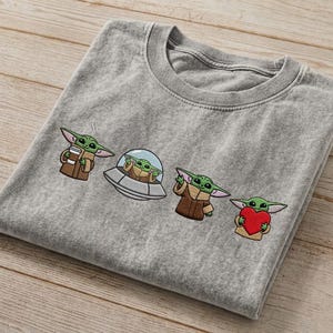 May include: A gray t-shirt featuring embroidered images of Grogu from The Mandalorian. The designs include Grogu holding a cup, in a spaceship, wearing a cloak, and holding a heart. The shirt is made of a soft material.