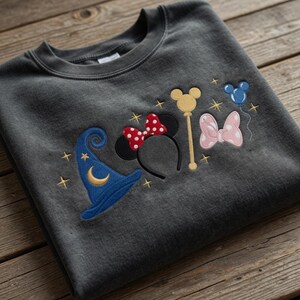 Pode incluir: Um moletom cinza escuro com desenhos bordados: um chap&eacute;u de mago azul, orelhas da Minnie com um la&ccedil;o vermelho de bolinhas, uma varinha dourada, um la&ccedil;o rosa de bolinhas e bal&otilde;es azuis. O design &eacute; adornado com estrelas douradas.