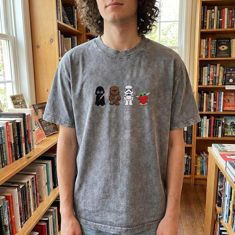 May include: Grey, short-sleeved t-shirt with a cartoon design of Star Wars characters: Darth Vader, Chewbacca, a Stormtrooper, and Baby Yoda holding a heart. The shirt has a vintage, washed-out appearance.