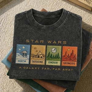 May include: Dark grey t-shirt with gold embroidered "STAR WARS" above four panels depicting scenes from the Star Wars universe. Panels show Hoth, Tatooine, Endor, and Geonosis. The phrase "A GALAXY FAR, FAR AWAY" is embroidered below. The t-shirt is folded on top of other t-shirts.