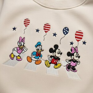 Puede incluir: Sudadera color crema con bordados de los personajes de Disney Daisy Duck, Donald Duck, Mickey Mouse y Minnie Mouse cruzando un paso de cebra. Globos y estrellas rojos, blancos y azules bordados.