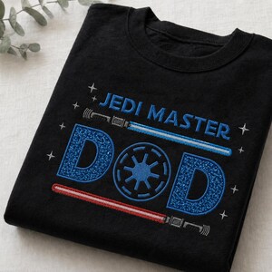 May include: Black t-shirt with the words "JEDI MASTER DAD" in blue and white embroidery. The design includes a blue lightsaber, a red lightsaber, and a blue emblem. The shirt is folded.