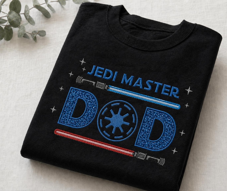 May include: Black t-shirt with the words "JEDI MASTER DAD" in blue and white embroidery. The design includes a blue lightsaber, a red lightsaber, and a blue emblem. The shirt is folded.