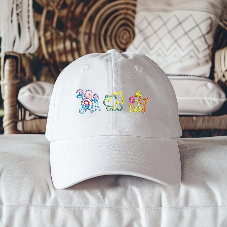 May include: A white baseball cap featuring three colorful embroidered cartoon characters. The cap has a curved brim and a classic six-panel design, suitable for casual wear. The embroidered designs are outlined in rainbow colors.