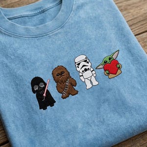 May include: Pale blue crew neck sweatshirt featuring embroidered Star Wars characters: Darth Vader, Chewbacca, a Stormtrooper, and Grogu holding a heart. The characters are aligned across the front.