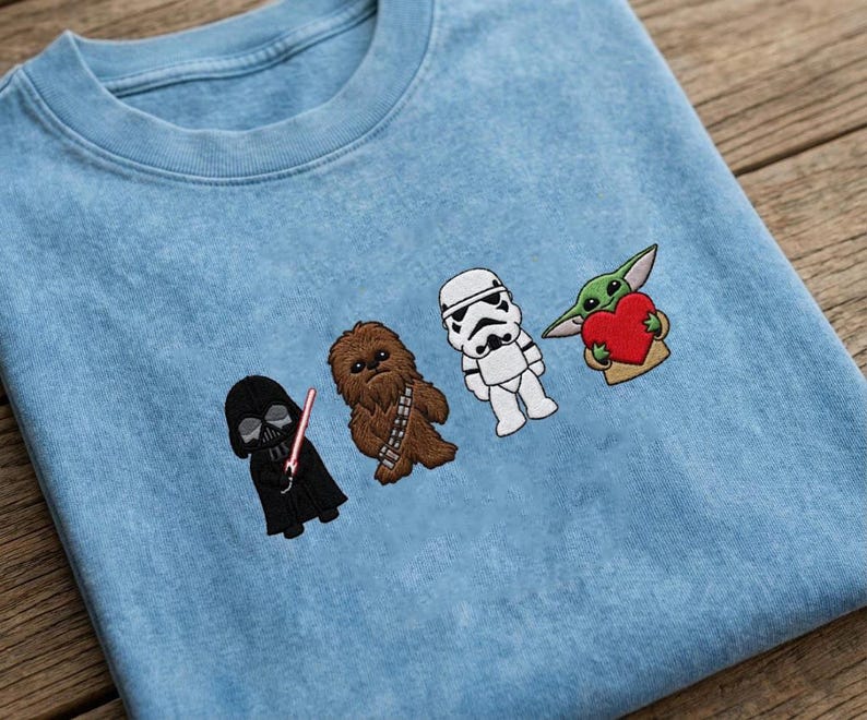 May include: Pale blue crew neck sweatshirt featuring embroidered Star Wars characters: Darth Vader, Chewbacca, a Stormtrooper, and Grogu holding a heart. The characters are aligned across the front.
