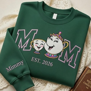 Puede incluir: Una sudadera verde oscuro con la palabra "MOM" en rosa, con un diseño de taza y tetera de dibujos animados. El texto "EST. 2026" está debajo del diseño, y la palabra "Mimmy" está en la manga.