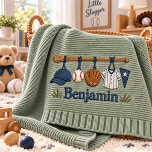 以下が含まれることがあります： セージグリーンのニット製ベビーブランケット。野球をテーマにした刺繍が施されています。木製のバットに、青いキャップ、野球ボール、茶色のグローブ、白いユニフォーム、青いペナントが吊り下げられたデザインです。青い糸で「Benjamin」という名前が刺繍されています。