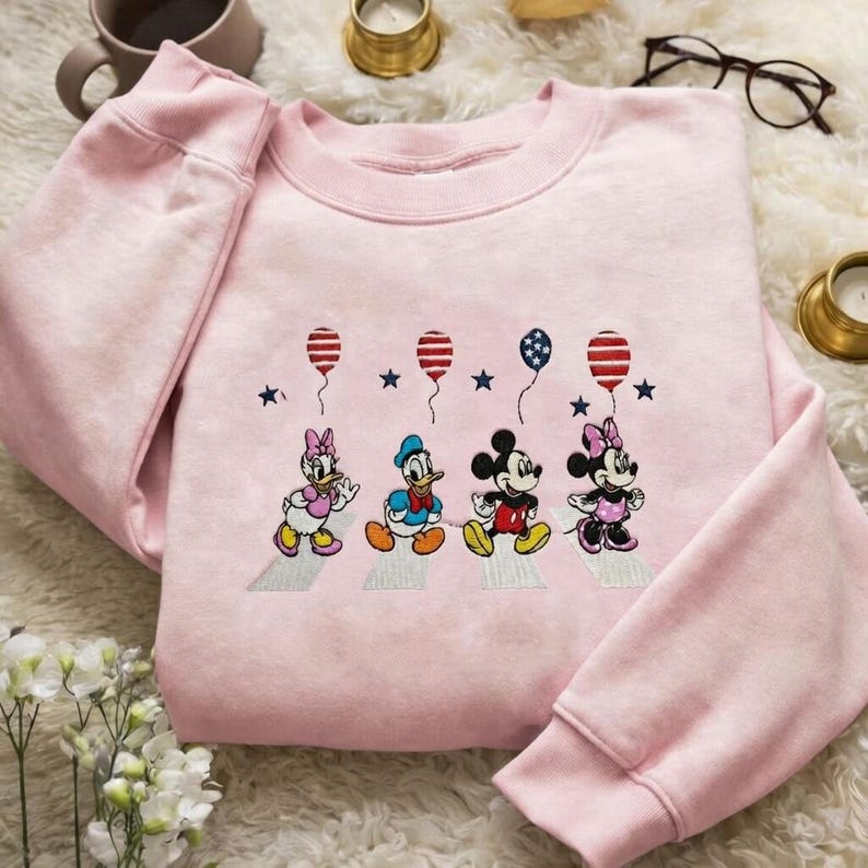 Puede incluir: Una sudadera rosa claro con un dise&ntilde;o bordado de los personajes de Disney Daisy Duck, Donald Duck, Mickey Mouse y Minnie Mouse caminando. Cada personaje sostiene un globo. El dise&ntilde;o incluye estrellas y globos rojos, blancos y azules.