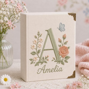 Könnte beinhalten: Ein cremefarbenes, stoffbezogenes Buch mit einem gestickten salbeigrünen Buchstaben A, dem Namen Amelia in Schreibschrift und Blumenmotiven in Rosa- und Grüntönen. Ein kleiner blauer Schmetterling ist über dem Buchstaben gestickt und die Ecken haben dekorative goldfarbene Metallbeschläge.