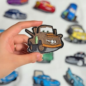 Könnte beinhalten: Gestickter Aufnäher mit der Figur Mater aus dem Film Cars. Der Aufnäher ist braun mit grünen und orangen Akzenten und zeigt ein lächelndes Gesicht. Weitere Autoaufnäher sind im Hintergrund zu sehen.