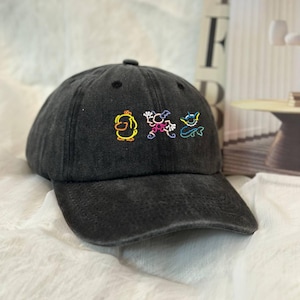 May include: A black baseball cap with a curved brim. The cap features colorful embroidered cartoon characters: a yellow duck, a pink creature, and a blue monster. The cap has a distressed, worn look.