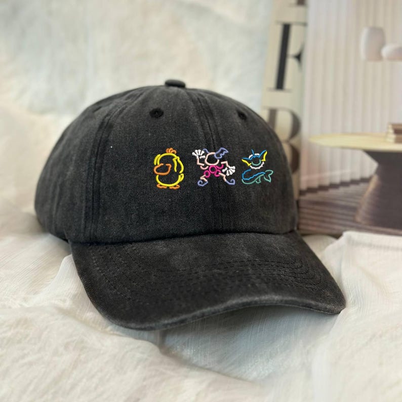 May include: A black baseball cap with a curved brim. The cap features colorful embroidered cartoon characters: a yellow duck, a pink creature, and a blue monster. The cap has a distressed, worn look.