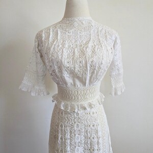 May include: A white eyelet dress with a high neckline and short sleeves. The dress has a fitted bodice and a full skirt. The dress is made of a delicate fabric with a lace trim.