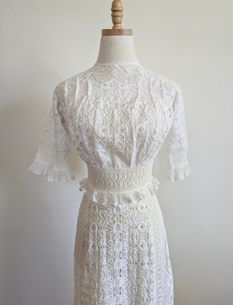 May include: A white eyelet dress with a high neckline and short sleeves. The dress has a fitted bodice and a full skirt. The dress is made of a delicate fabric with a lace trim.