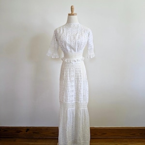 May include: A white, floor-length, eyelet dress with a high neck and short sleeves. The dress has a fitted bodice and a flowing skirt with multiple tiers.