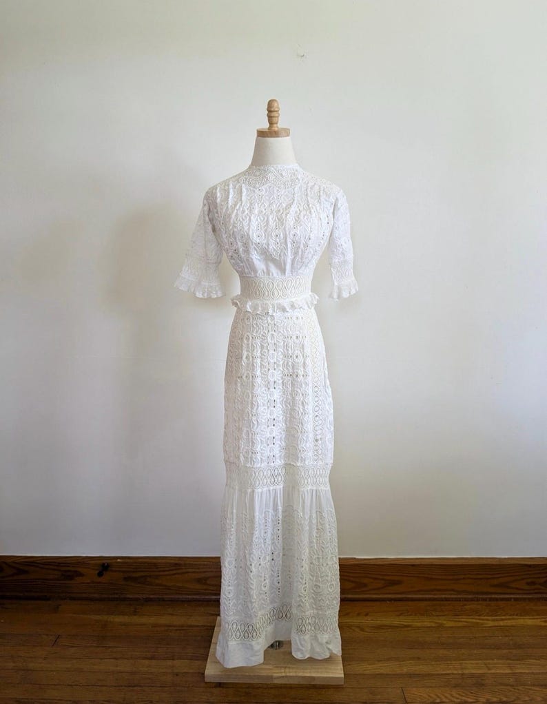 May include: A white, floor-length, eyelet dress with a high neck and short sleeves. The dress has a fitted bodice and a flowing skirt with multiple tiers.