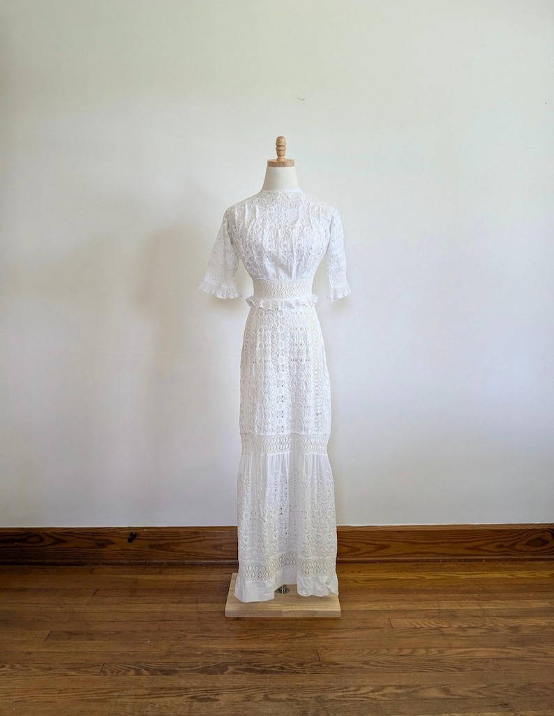 May include: A white eyelet maxi dress on a dress form. The dress has short sleeves, a fitted bodice, and a tiered skirt. The fabric is delicate and lacy. The dress is a vintage style, perfect for a wedding or special occasion. The dress is shown against a white wall and wooden floor.