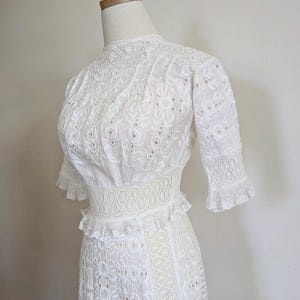 May include: A white eyelet dress with a high neck, short sleeves, and a fitted waist. The dress has a delicate floral pattern and a ruffled skirt.