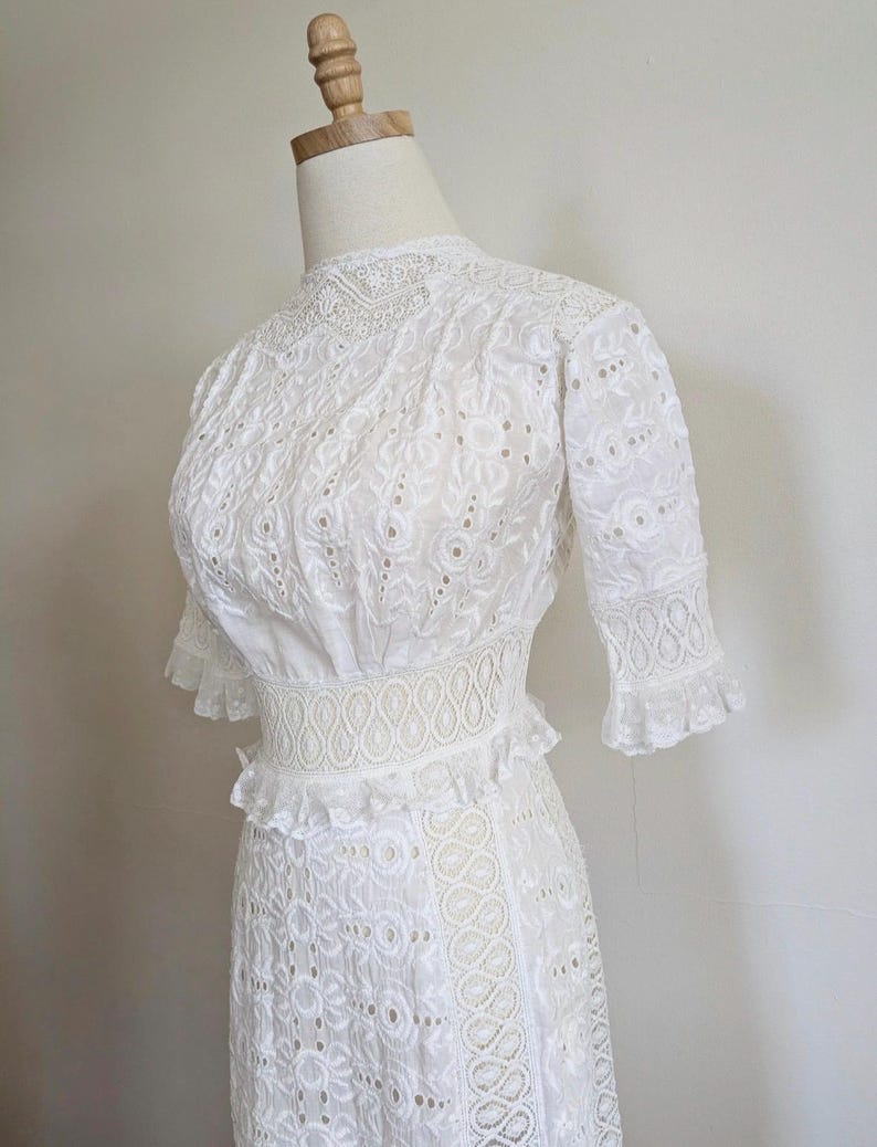 May include: A white eyelet dress with a high neck, short sleeves, and a fitted waist. The dress has a delicate floral pattern and a ruffled skirt.