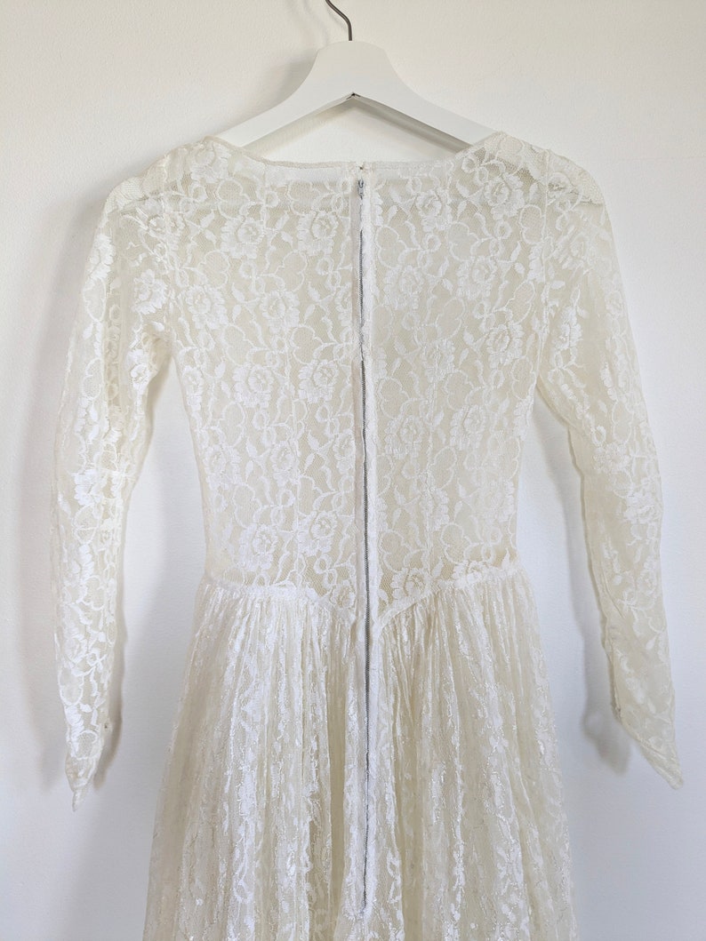 Puede incluir: Un vestido blanco de encaje con mangas largas y una cremallera en la espalda. El vestido tiene un corpi&ntilde;o ajustado y una falda fluida.