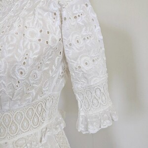 May include: White lace dress with a high neck and short sleeves. The dress has a delicate floral pattern and a ruffled hem.