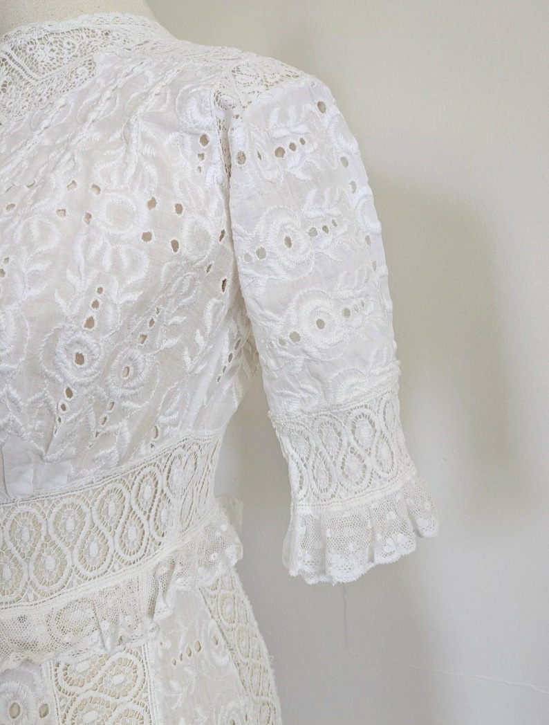 May include: White lace dress with a high neck and short sleeves. The dress has a delicate floral pattern and a ruffled hem.
