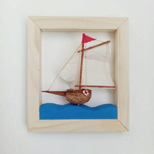 May include: A small wooden sailboat with a white sail and a red flag, floating on blue waves, framed in a natural wood frame.