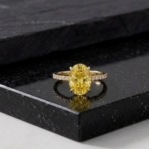 May include: A gold ring featuring a large, oval-cut yellow gemstone. The band is embellished with smaller, clear gemstones. The ring is displayed on a dark, reflective surface, accentuating its sparkle and design.
