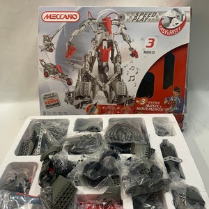 May include: A Meccano Speed Play construction set with three models, including a robot with music and movements. The box shows the robot in the center, with a crane and a car on either side. The box is red and gray with the words "Speed Play" and "Easy & Fast!" printed on it. The box also includes a picture of a child building the robot.