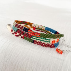 Könnte beinhalten: Ein mehrfarbiges Perlenarmband mit verschiedenen Mustern in Rot, Grün, Blau und Weiß. Es hat eine silberne Kette und einen kleinen orangefarbenen Blumenanhänger. Das Armband liegt auf einer weißen Oberfläche.
