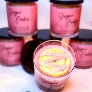 Peut inclure: Cinq pots de bougies en verre rose avec des couvercles noirs. Les pots de bougies sont &eacute;tiquet&eacute;s "Sugar Cookie" et ont un biscuit au sucre rose sur le dessus de la bougie dans le pot du centre.